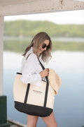 Woman holding a large beige and black tote bag with 'Muskoka Always' text by a waterfront.