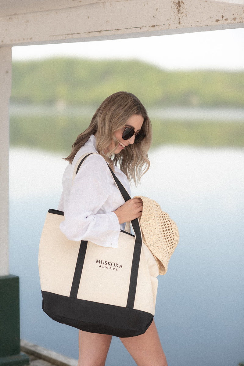 Woman holding a large beige and black tote bag with 'Muskoka Always' text by a waterfront.