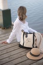 Woman sitting on a dock with a large beige and black tote bag and hat, near a lake.