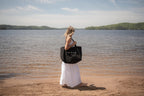Woman holding a black 'lake it easy' tote bag by a lake