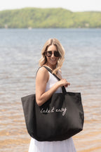 Woman holding a black tote bag with 'lake it easy' text by a lake.