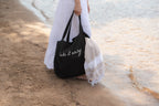 Person holding a black tote bag with 'lake it easy' text on a beach.