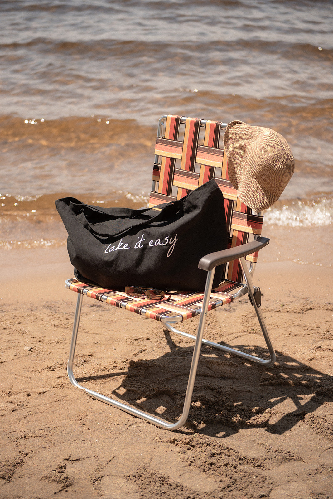 Beach chair with a patterned backrest and 'lake it easy' bag on a sandy beach.