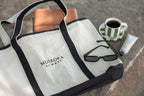 White and black tote bag with 'Muskoka Always' branding, sunglasses, and a coffee cup on a stone surface.