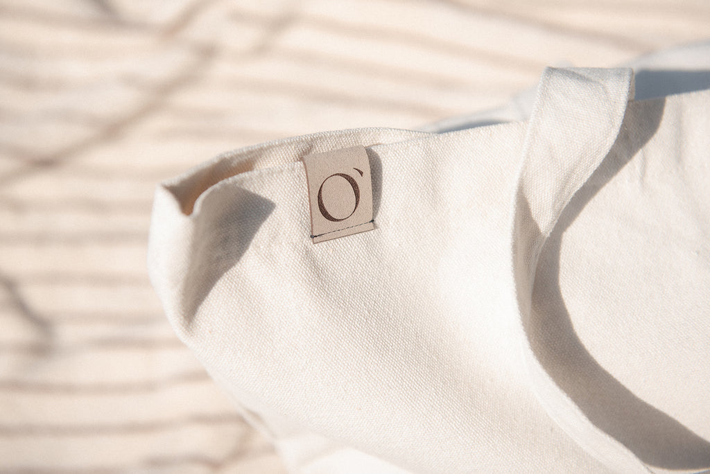 Close-up of a light beige canvas tote bag with a small square leather patch featuring a stylized 'O' logo, set against a blurred background with soft shadows.
