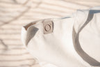 Close-up of a light beige canvas tote bag with a small square leather patch featuring a stylized 'O' logo, set against a blurred background with soft shadows.