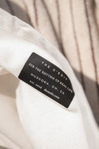 Close-up of a black fabric brand label sewn onto a light cream-colored tote bag. The label reads 'THE O'EDIT FOR THE RHYTHM OF REAL LIFE MUSKOKA, ON, CA'.
