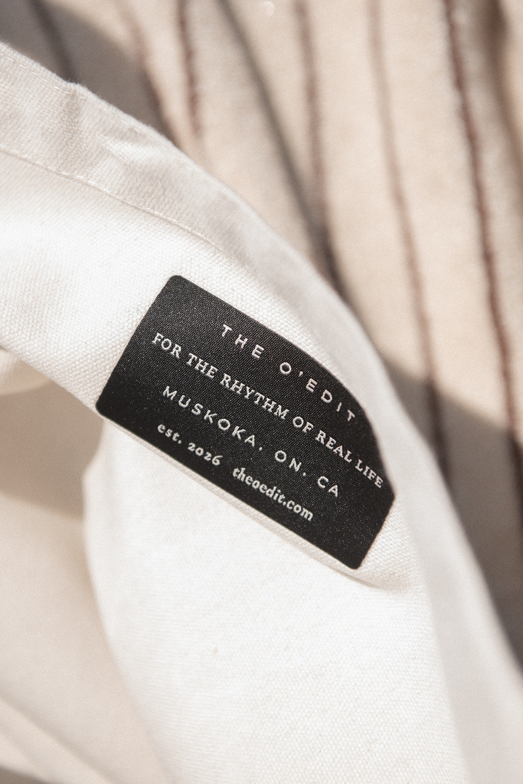 Close-up of a black fabric brand label sewn onto a light cream-colored tote bag. The label reads 'THE O'EDIT FOR THE RHYTHM OF REAL LIFE MUSKOKA, ON, CA'.