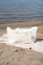 A white rectangular beach bag with 'lake it easy' embroidered in gold script, resting on a striped beach towel on the sand near the lake.