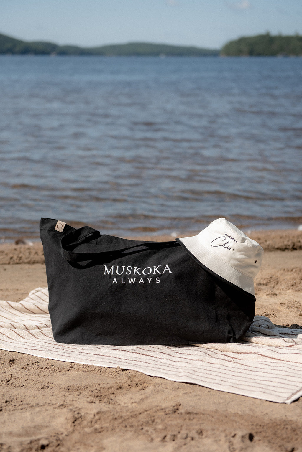 A black tote bag with 'MUSKOKA ALWAYS' printed in white sits on a sandy beach next to a striped towel, with a white sun hat and a scenic lake in the background.
