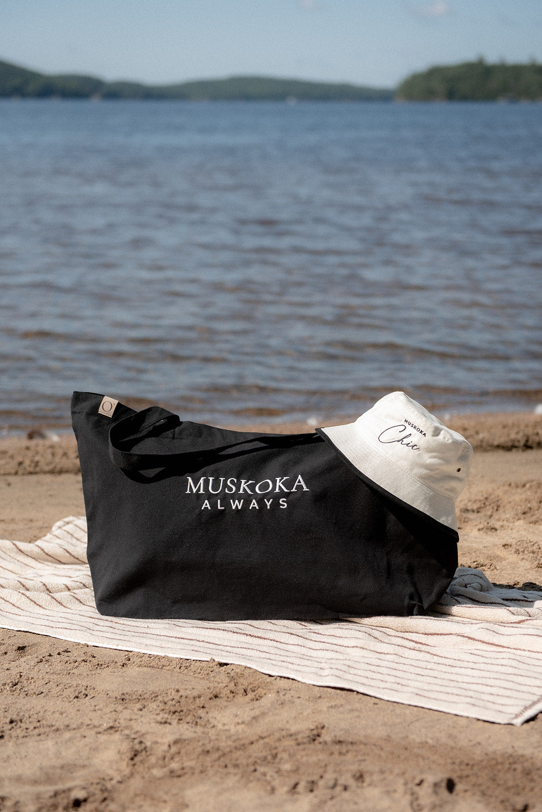 A black tote bag with 'MUSKOKA ALWAYS' printed in white sits on a sandy beach next to a striped towel, with a white sun hat and a scenic lake in the background.