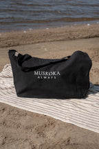 A black tote bag with 'MUSKOKA ALWAYS' printed in white text, resting on a striped beach towel on a sandy beach with the lake in the background.
