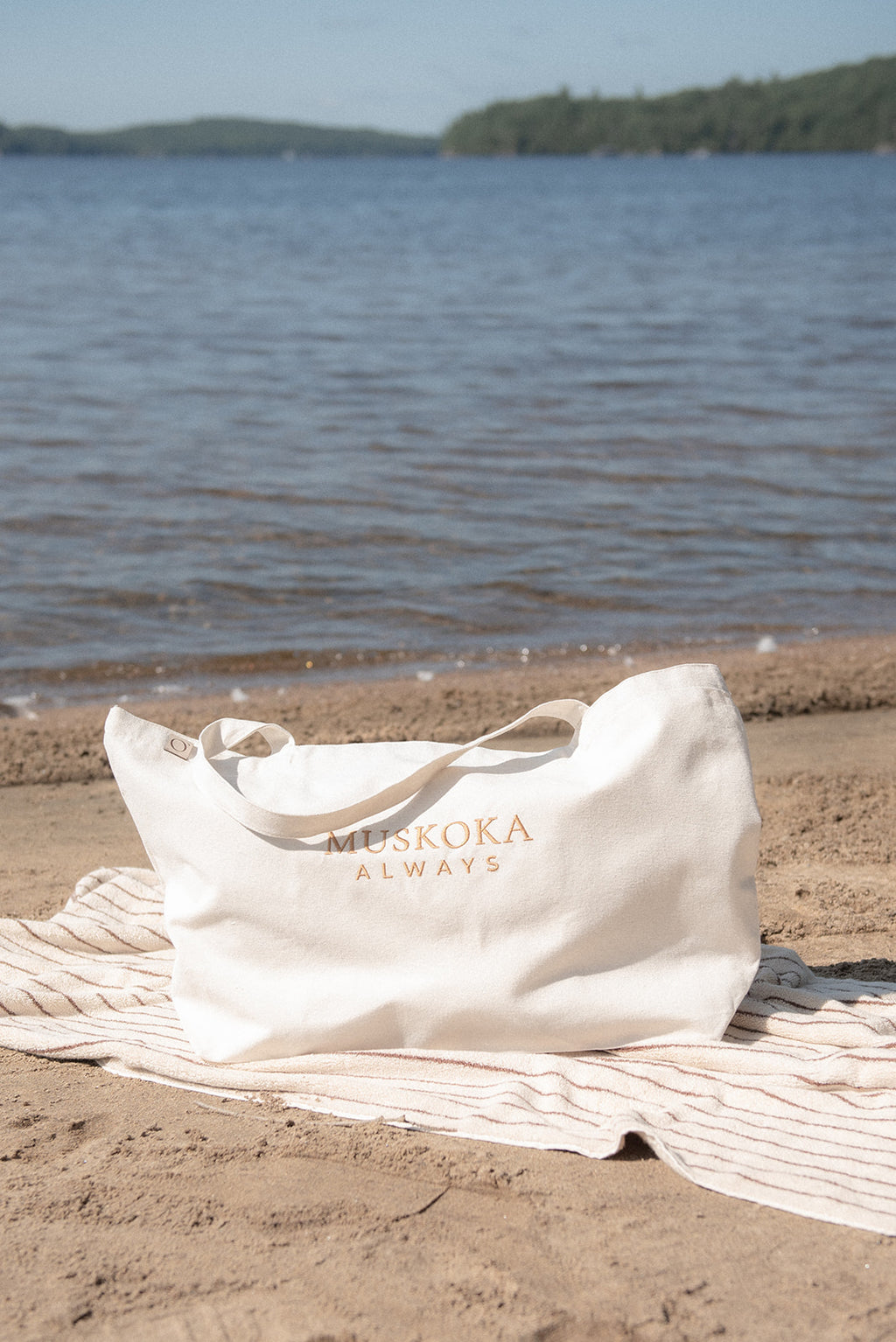 An off-white tote bag with 'MUSKOKA ALWAYS' printed in gold lettering sits on a striped beach towel on the sand, with a lake and forested shoreline in the background.