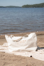 An off-white tote bag with 'MUSKOKA ALWAYS' printed in gold lettering sits on a striped beach towel on the sand, with a lake and forested shoreline in the background.