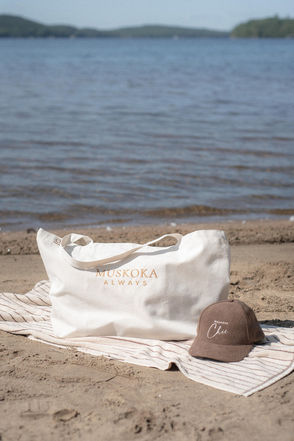 A white tote bag with 'MUSKOKA ALWAYS' printed in gold on a beach towel next to a brown baseball cap, set against a blurred lake background.