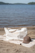 A white tote bag with 'MUSKOKA ALWAYS' printed in gold on a beach towel next to a brown baseball cap, set against a blurred lake background.