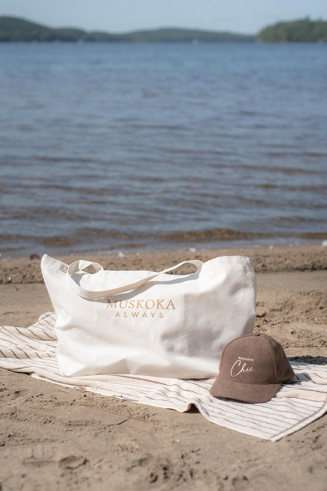 A white tote bag with 'MUSKOKA ALWAYS' printed in gold on a beach towel next to a brown baseball cap, set against a blurred lake background.