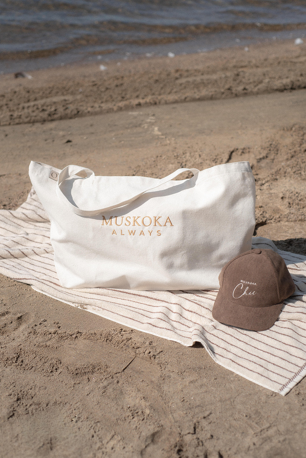 A white tote bag with 'MUSKOKA ALWAYS' printed in gold lettering, resting on a striped beach towel on a sandy beach next to a brown baseball cap.