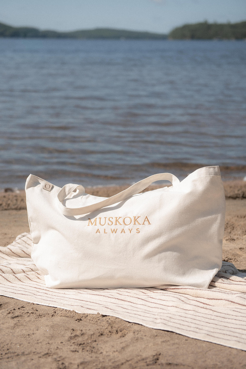 An off-white tote bag with gold lettering that reads 'MUSKOKA ALWAYS' sits on a striped beach towel on the sand, with a lake and distant shoreline in the background.