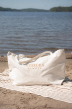 An off-white tote bag with gold lettering that reads 'MUSKOKA ALWAYS' sits on a striped beach towel on the sand, with a lake and distant shoreline in the background.