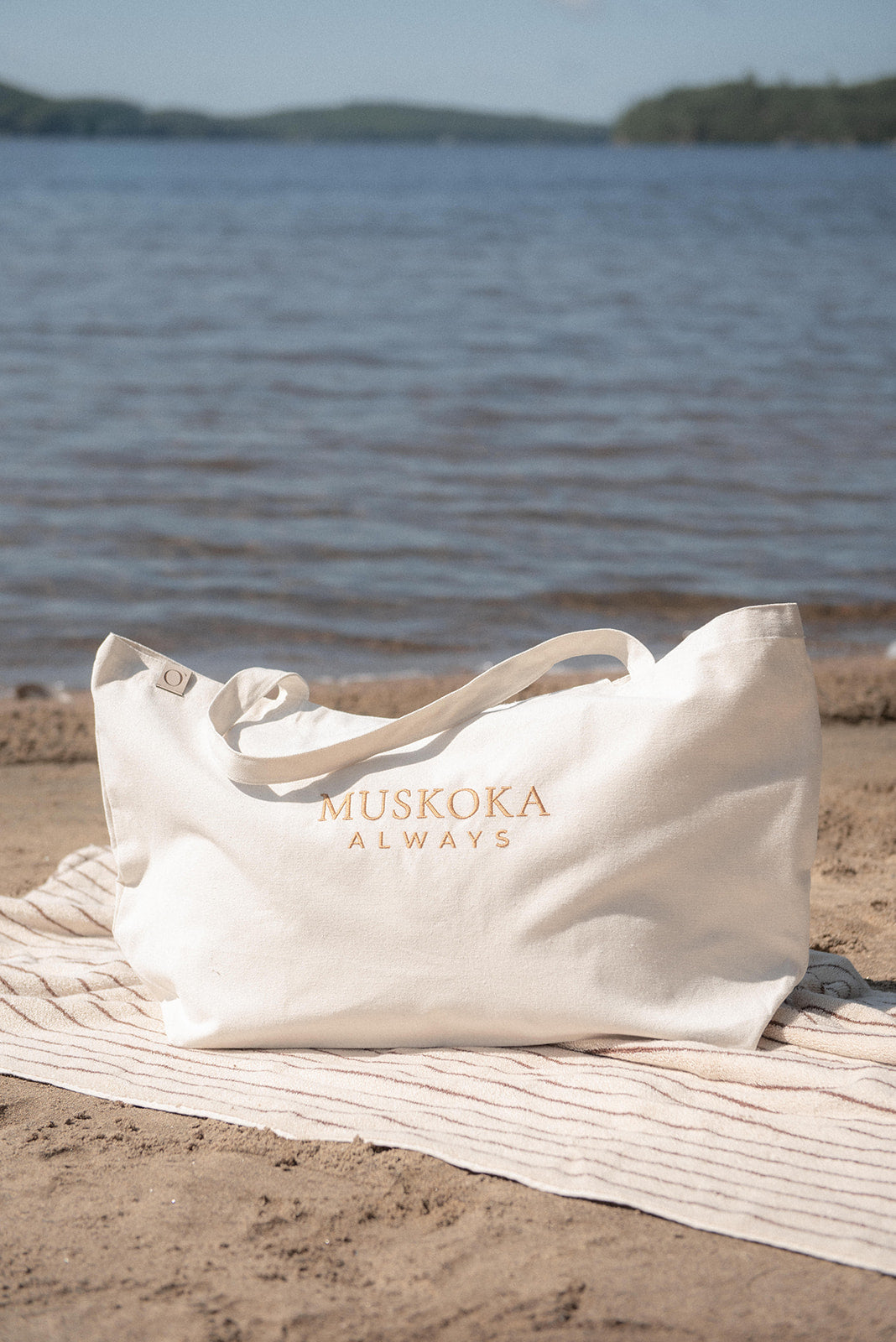 An off-white tote bag with gold lettering that reads 'MUSKOKA ALWAYS' sits on a striped beach towel on the sand, with a lake and distant shoreline in the background.